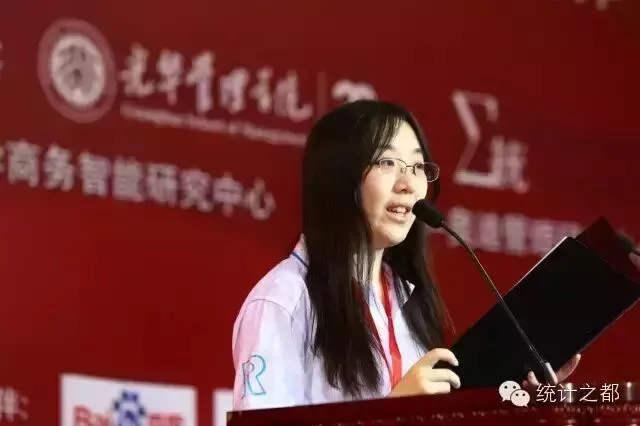 一名身穿白色会务衫的发言者站在讲台前致辞，身后是红色会议背景板。
