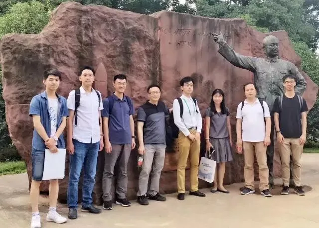 九名参会者在校园户外的一座人物雕像前并排合影。