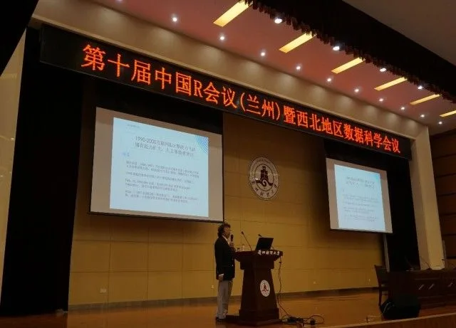 宽阔礼堂内，一名演讲者站在双投影屏幕之间的讲台前。