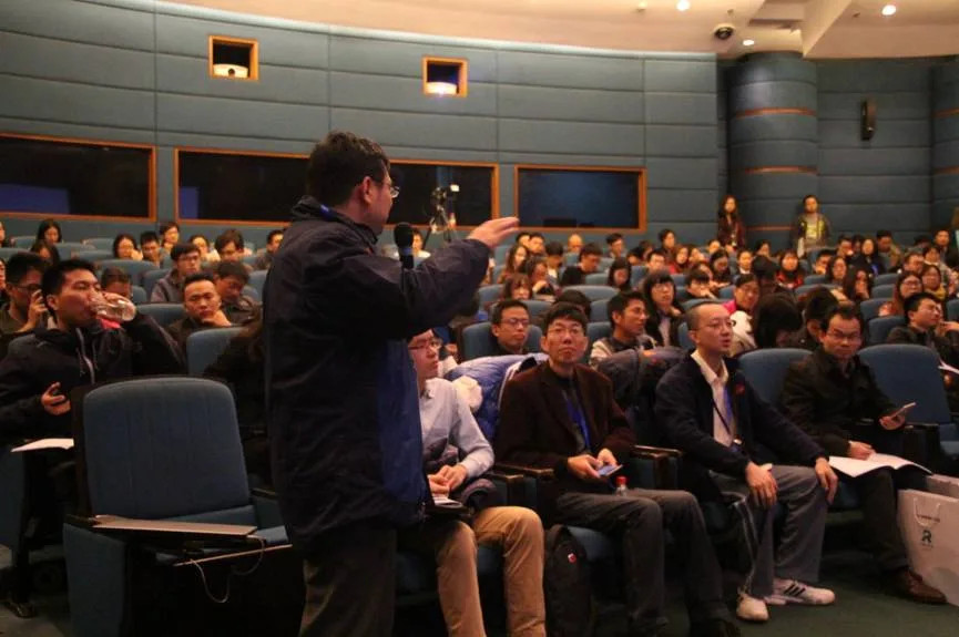 一名参会者站在座位间举手发言，周围听众转身看向他。