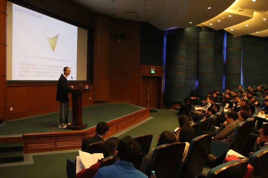 演讲者站在投影屏幕前发言，阶梯座席中的听众面向讲台。