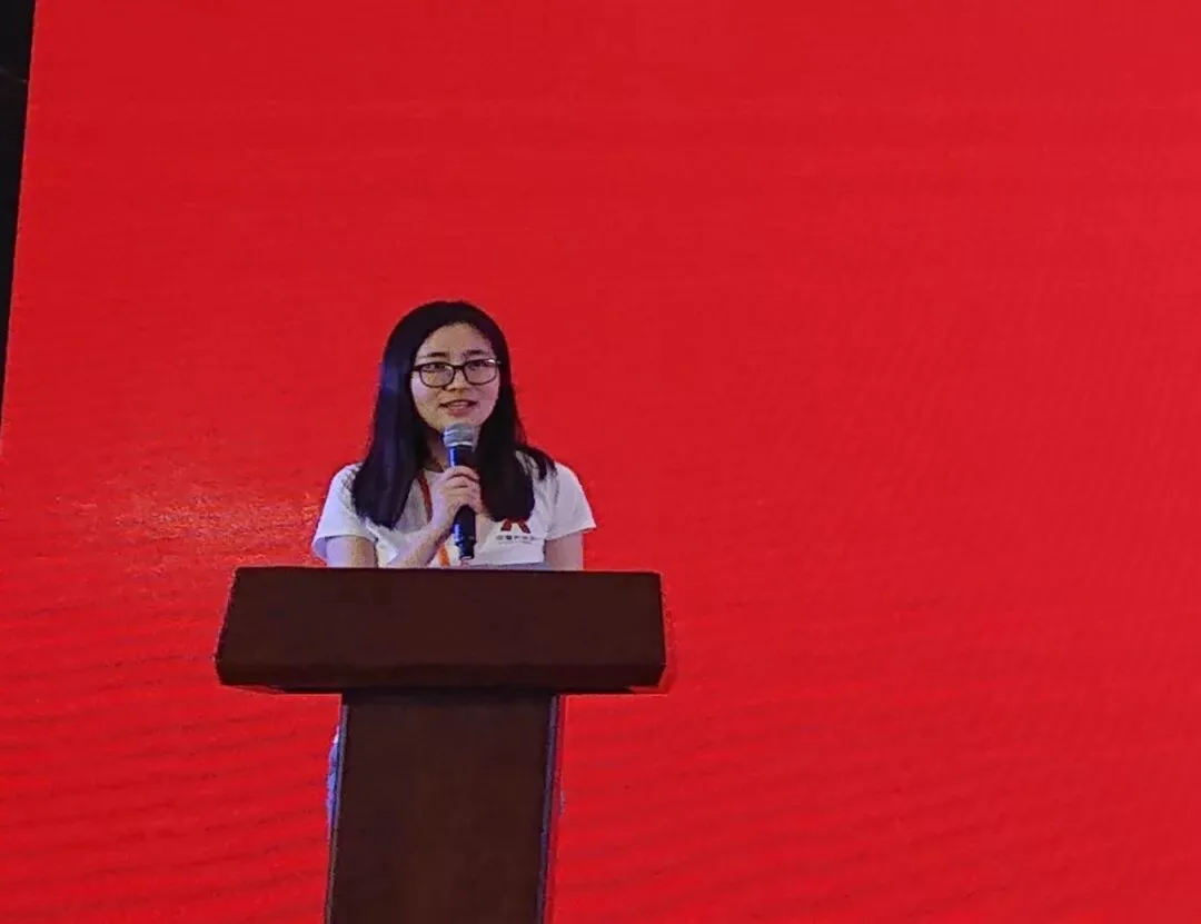 一名身穿白色会务衫的发言者在红色舞台背景前致辞。