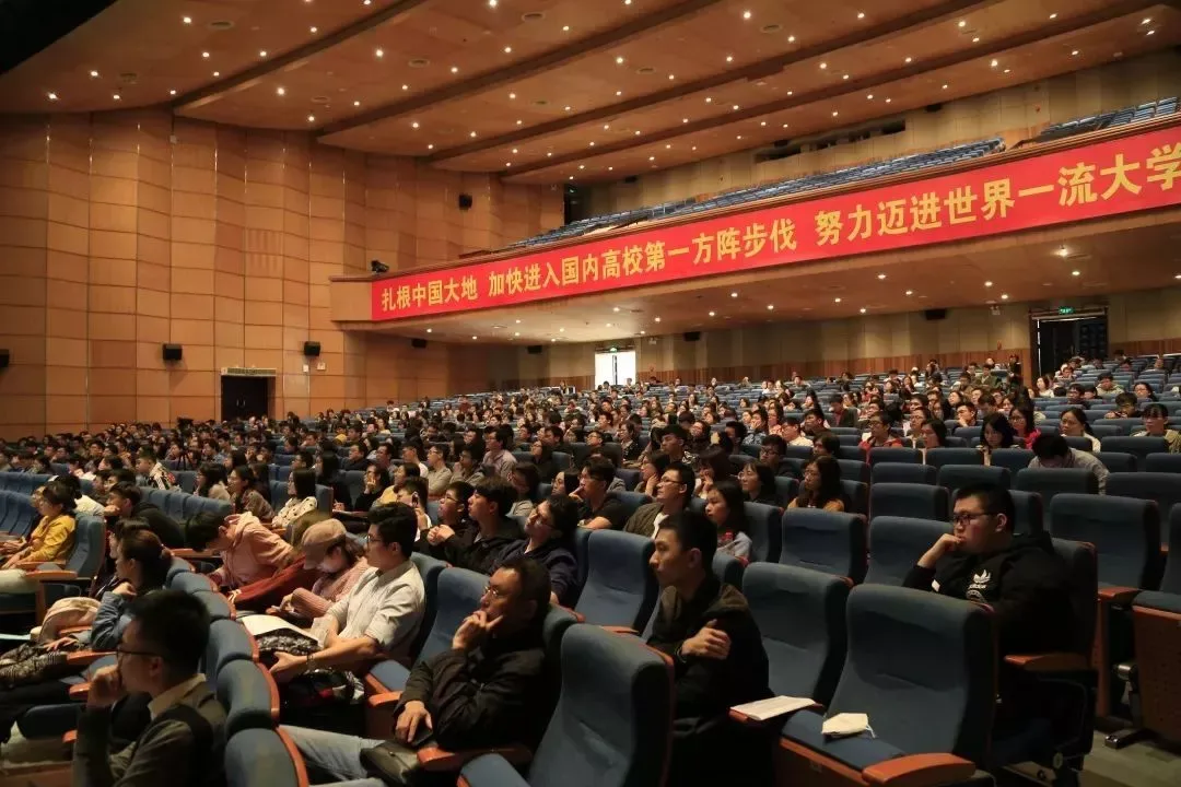 大型礼堂内坐满面向舞台的听众，红色横幅悬挂在会场上方。