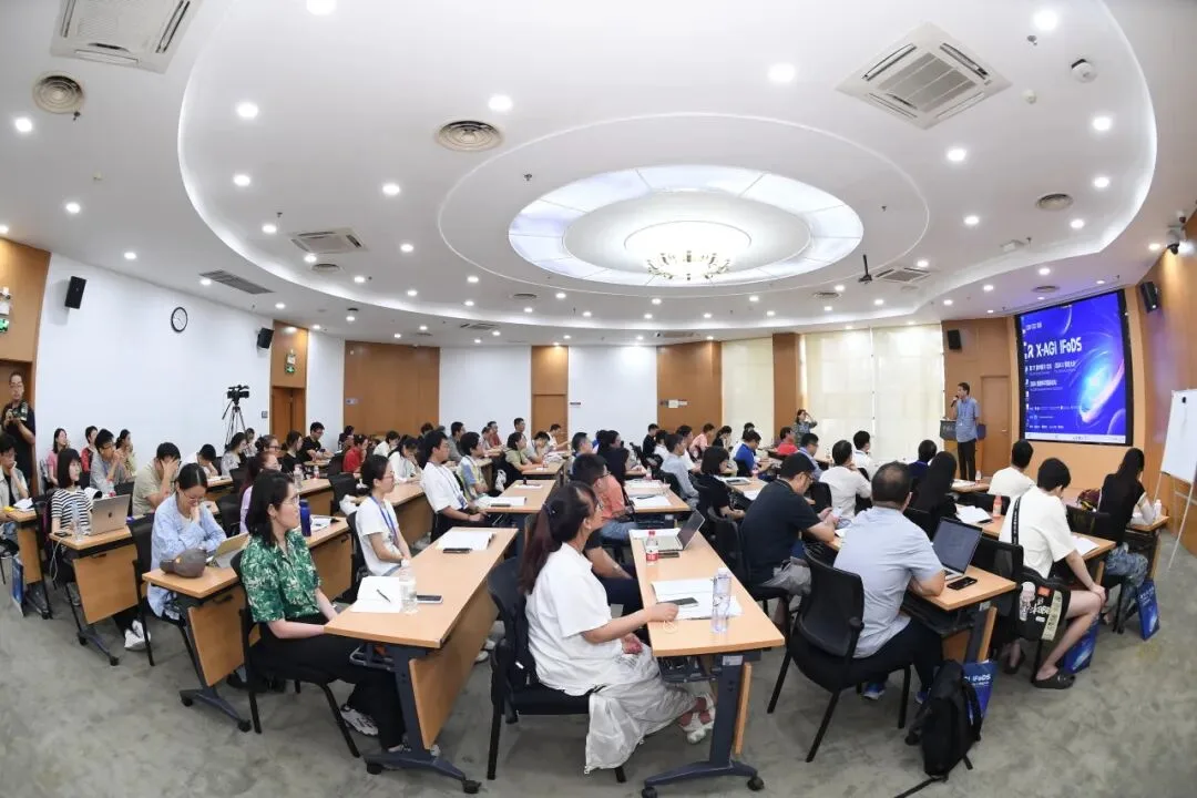 明亮的会议室内坐满参会者，前方发言者站在显示X-AGI与IFoDS会议信息的屏幕旁。