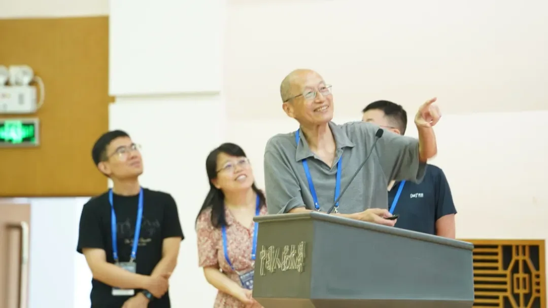 吴喜之教授在中国人民大学讲台后微笑致辞，身旁几位老师一同望向会场。