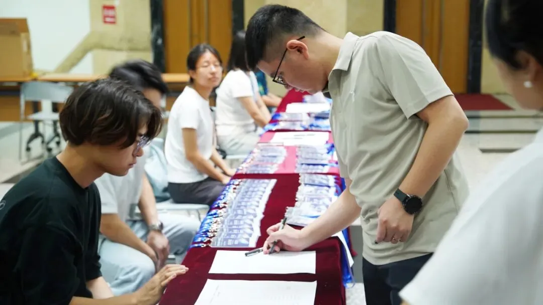 参会者俯身在签到表上填写信息，桌后会务人员旁摆放着证件与会议材料。