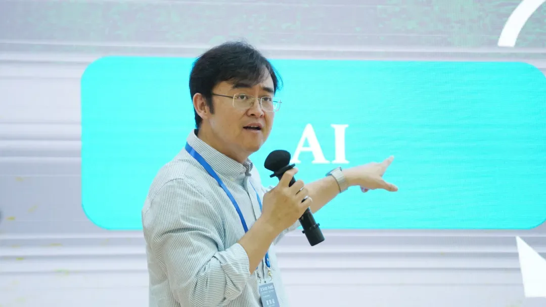 一名演讲者手持话筒指向身后屏幕上的AI字样，屏幕为青绿色色块。