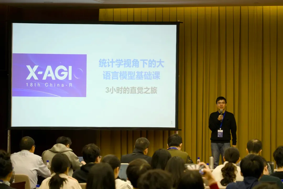 演讲者站在大型投影屏幕旁，屏幕显示统计学视角下的大语言模型基础课标题，前方坐有学员。