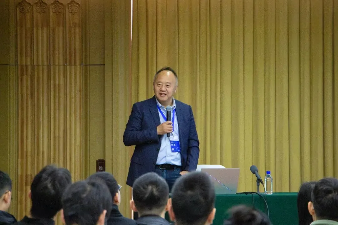 一名手持话筒的演讲者站在会场前方发言，前景可见多排听众。